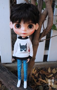 a doll with black hair wearing a sweater and jeans standing next to a tree in front of a white picket fence