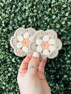 two crocheted flowers are held up in front of a green bush with leaves