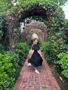 a woman in a black dress is walking down a brick path surrounded by greenery