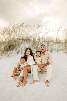 a family sitting in the sand at the beach with tall grass and sky behind them