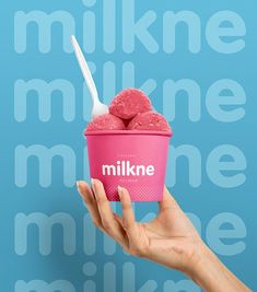 a hand holding up a pink ice cream cup with a spoon in it, against a blue background