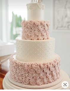 a three tiered cake with white frosting and pink roses on top, sitting on a table