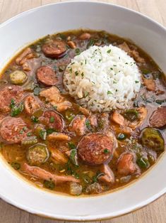 a white bowl filled with gumbo and sausage stew next to rice on a wooden table