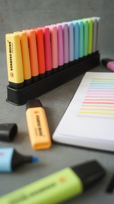 there are many different colored pens on the table next to each other and a notepad