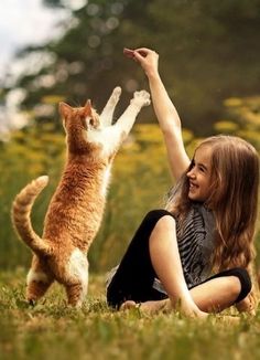 a woman sitting on the grass with her cat reaching up to catch something in the air