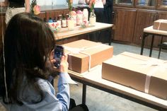 a woman sitting at a table with a cell phone in her hand and boxes on the counter behind her