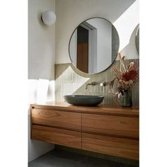 a bathroom sink sitting under a round mirror on top of a wooden counter next to a vase with flowers