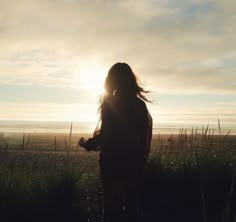 a woman standing on top of a grass covered field next to the ocean at sunset