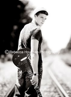 a black and white photo of a young man holding a guitar in front of train tracks