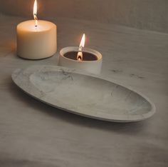 two lit candles sitting on top of a white plate next to a marble tray with a candle in it