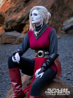 a woman with white makeup sitting on top of a rock wearing black and red clothing