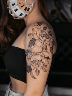 a woman with a mask on her face and flowers tattooed on her arm, behind her is a black and white photo