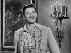 a man in a vest smiles as he stands next to a chandelier with candles on it