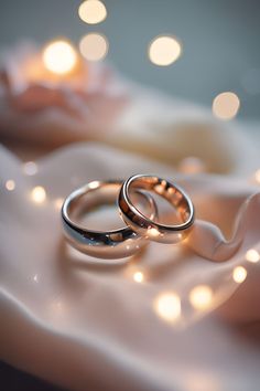 two wedding rings sitting on top of a white cloth covered tablecloth with lights in the background