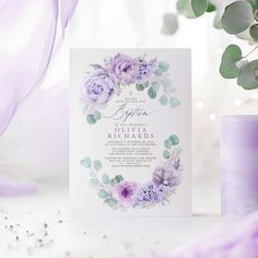 a purple and white wedding card with flowers on it next to a candle, ribbon and some greenery