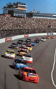 a group of cars driving down a race track in front of a large crowd on the stands