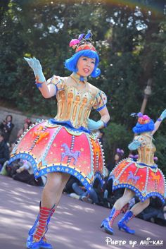 two women in colorful costumes are dancing on the street with trees and people behind them