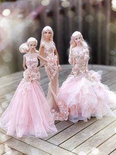 three barbie dolls in pink dresses standing on a wooden table with white lights behind them