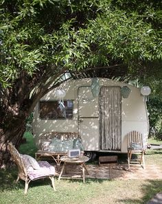 an old camper is parked under a tree with chairs and tables in the shade