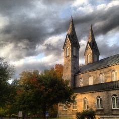 an old building with two towers under a cloudy sky and trees in the foreground