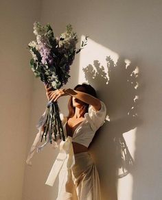 a woman holding flowers in her hands and leaning against a wall with the sun shining on her