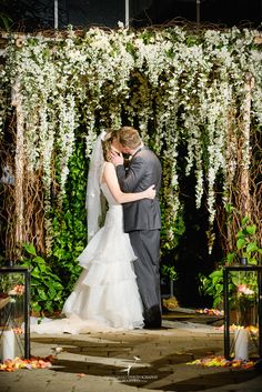 a bride and groom kissing in front of an outdoor wedding ceremony arch with white flowers