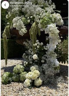 white flowers and greenery are arranged on the gravel in front of a garden fountain