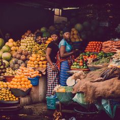 two women standing in front of a fruit stand with oranges, melons and other fruits