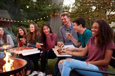 a group of young people sitting around a fire pit eating pizza and drinking sodas