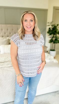 a woman standing in front of a bed wearing blue jeans and a white top with crochet on it