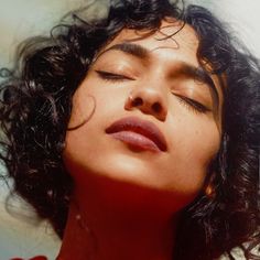 a woman with her eyes closed wearing a red shirt and black curly hair is looking up at the sky