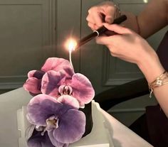 a woman lighting a candle on top of a cake with purple flowers and greenery