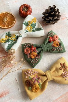 four bow ties with embroidered flowers and pinecones on them next to an orange slice