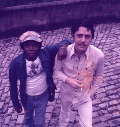 two young men standing next to each other on a cobblestone street with trees in the background