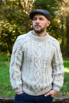 a man wearing a sweater and hat standing in front of some trees with his hands on his hips