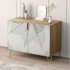 a white and brown cabinet with leaves on it next to a vase, potted plant and framed photograph