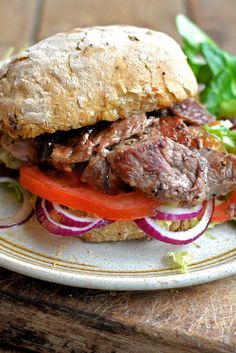 a sandwich with meat, tomatoes and onions on a white plate sitting on a wooden table