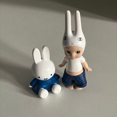 two toy rabbits sitting next to each other on a white counter top, one is wearing a bunny hat and the other has a blue shirt