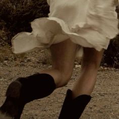 a woman in white dress and black boots running on dirt ground with trees in the background