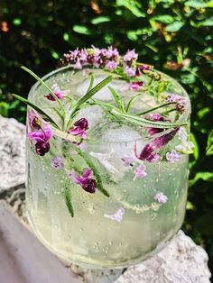 a glass filled with water and flowers sitting on top of a stone slab next to bushes