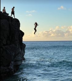 two people jumping off rocks into the ocean from a cliff above them, while another person watches