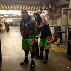 two men standing next to each other wearing green shorts and black shirts with tattoos on their arms