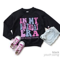 a black sweatshirt with pink and blue lettering on it, two pairs of sneakers are next to the sweater