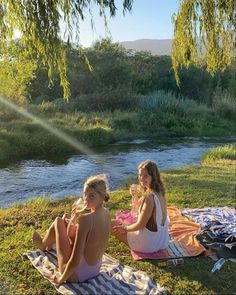 two young women sitting on blankets next to a river in the sun with trees and grass behind them