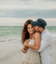 a man and woman hugging on the beach in front of the ocean while wearing hats