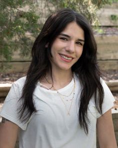 a woman with long hair standing in front of a wooden fence and smiling at the camera