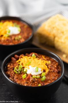 two black bowls filled with chili and sour cream on top of a table next to bread