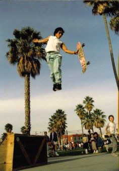 a young man jumping in the air with a skateboard near palm trees and other people