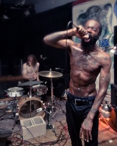 a shirtless man singing into a microphone while standing in front of a drum set