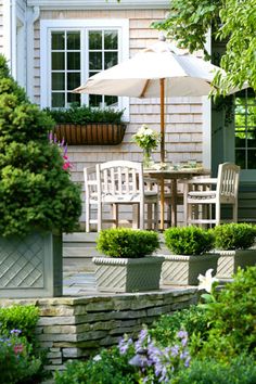 an outdoor patio with table and chairs surrounded by plants, shrubs and flowers in front of a house